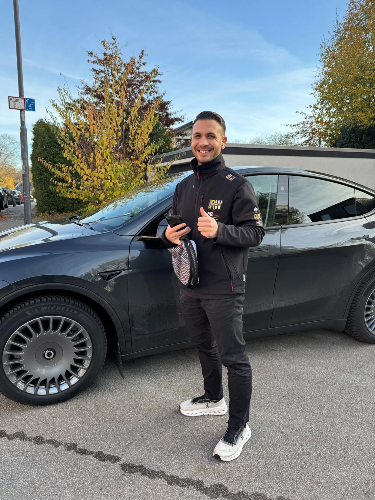 Ein junger Mann mit schwarzer Jacke lacht freundlich vor einem dunklen Auto