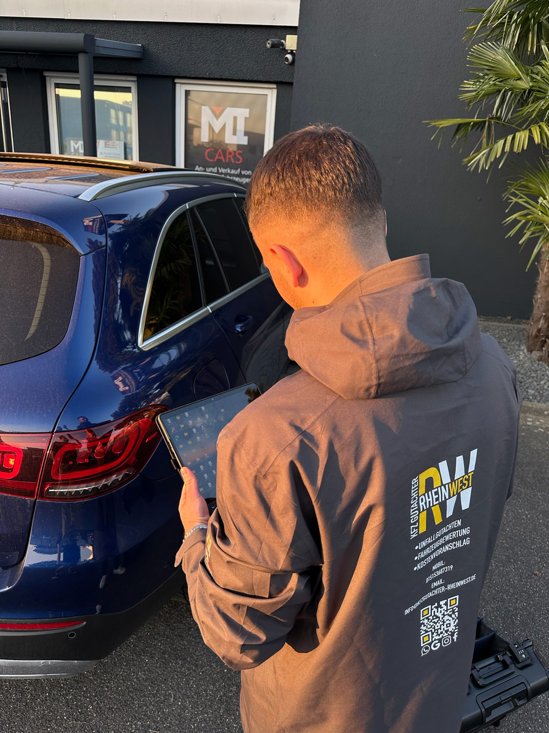 Ein Mann mit brauner Kurzhaarfrisur betrachtet konzentriert ein Tablet neben einem blauen Auto