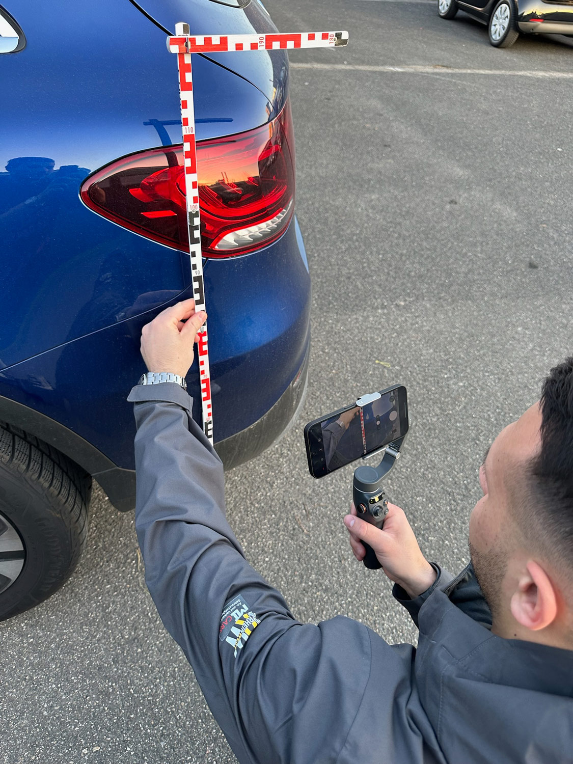 Ein Mann hält ein Smartphone auf einen blauen Wagen gerichtet und misst den Lackschaden mit einem rotweißen Messstab