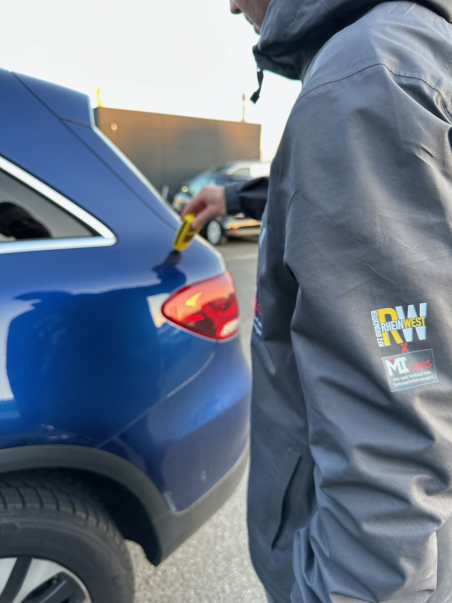 Eine Person im grauen Mantel misst fröhlich an einem blauen Auto mit gelbem Gerät