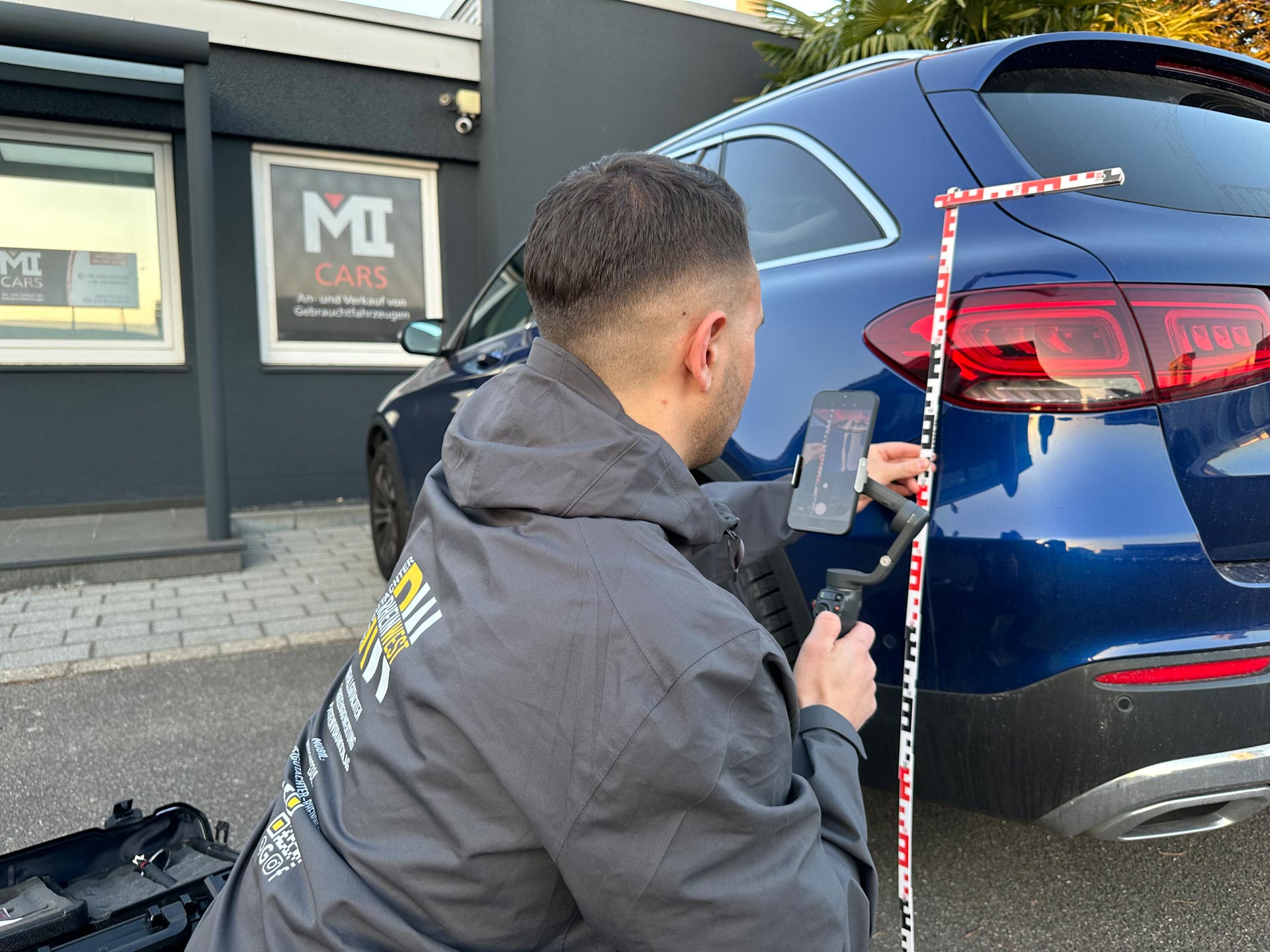 Ein Mann mit kurzem dunklem Haar misst an einem blauen Auto mit Maßband und Smartphone
