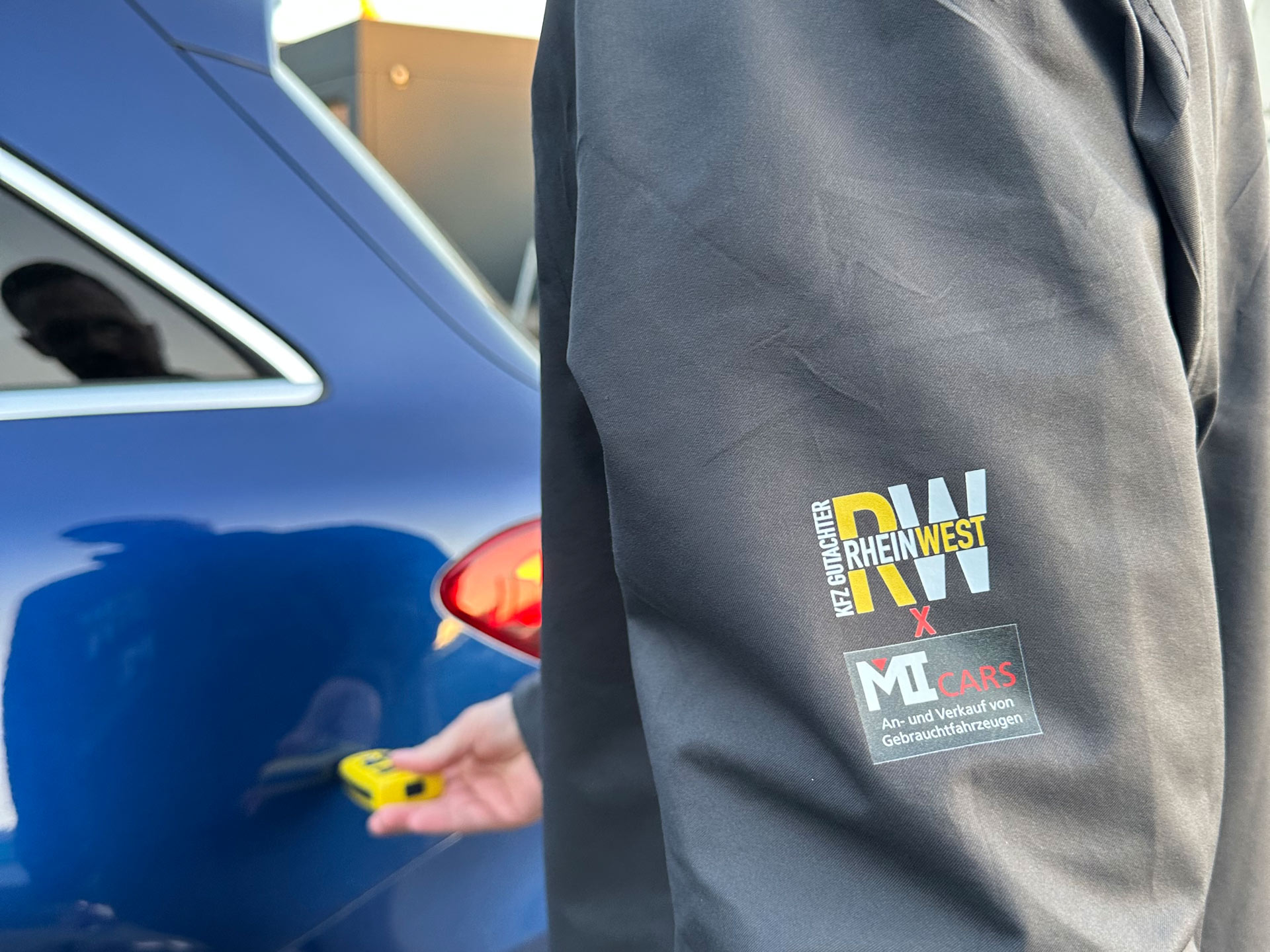 Eine Person mit dunkler Jacke misst mit einem gelben Gerät an einem blauen Auto