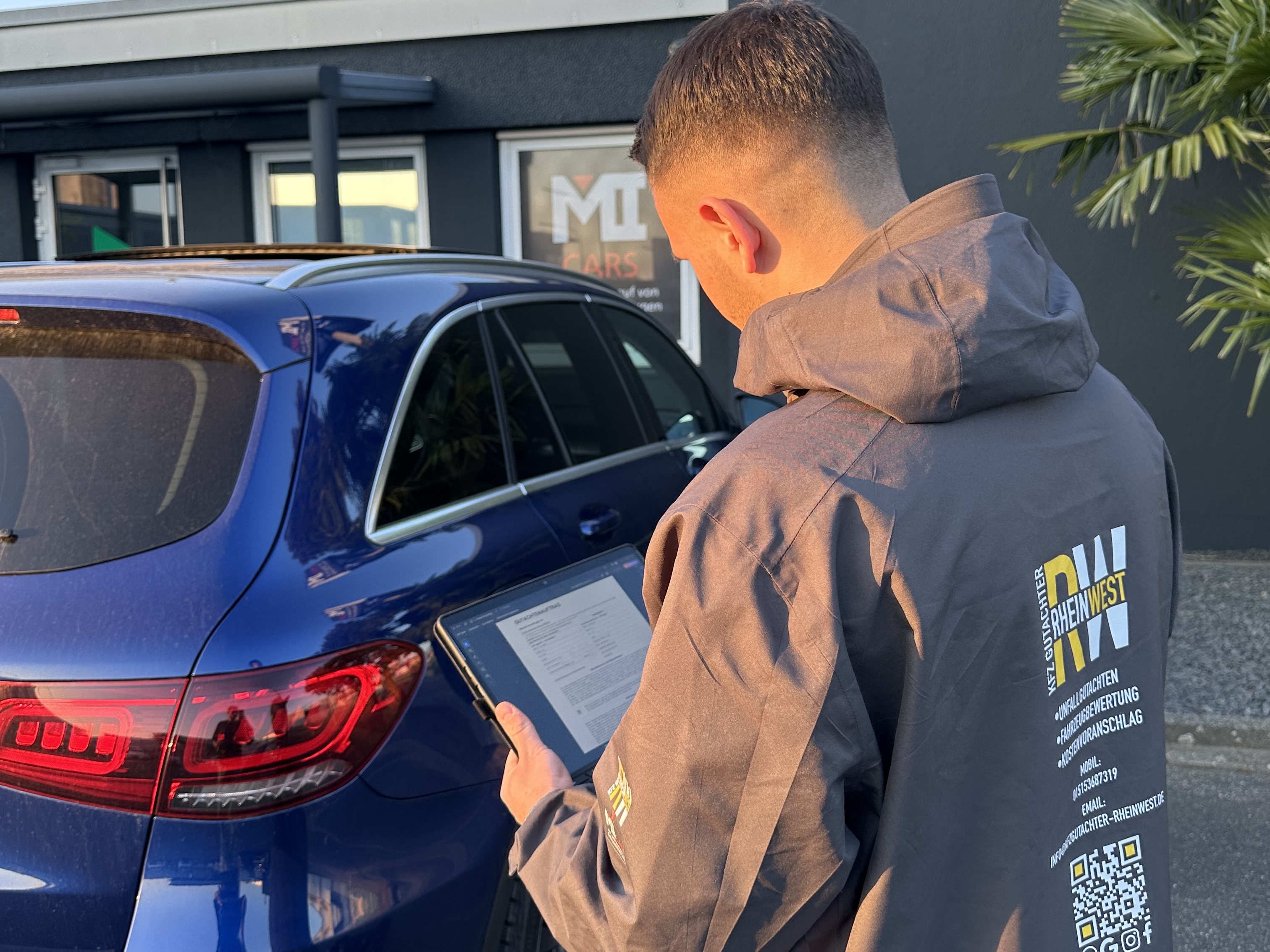 Ein junger Mann mit kurzem Haar betrachtet konzentriert ein Tablet neben einem blauen Auto