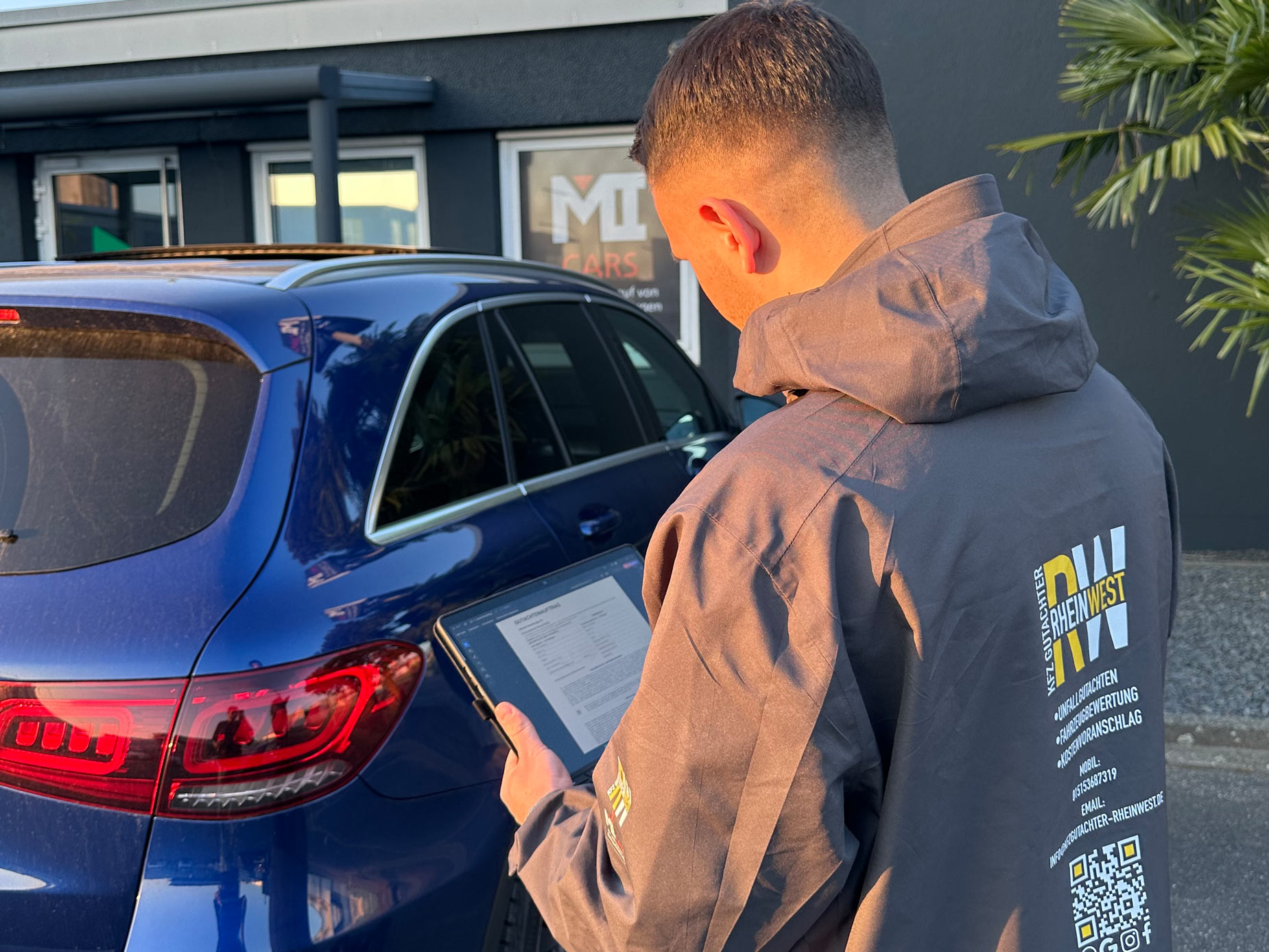 Ein Mann mit Kurzhaarschnitt steht draußen neben einem blauen Auto und liest ein Tablet