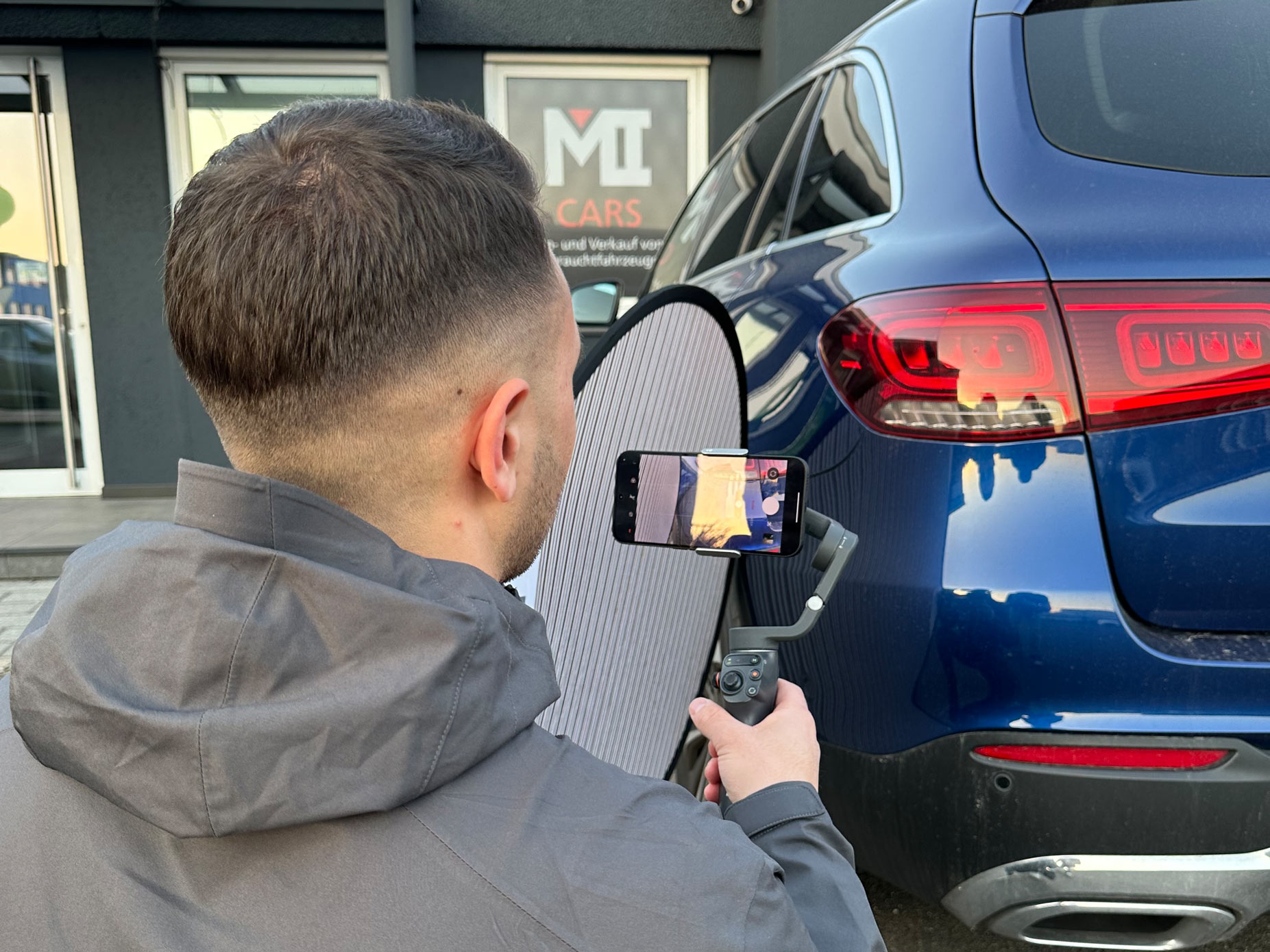 Ein Mann mit dunklen Haaren filmt lächelnd ein blaues Auto mit seinem Smartphone